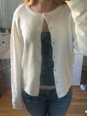 Vit basic kofta med rund hals - Enkel vit kofta med rund halsringning och knäppning framtill. Långärmad och mjuk, perfekt att slänga över ett linne eller t-shirt. Ribbstickade muddar vid ärmslut och nederkant. Passar till jeans eller kjol för en clean look.