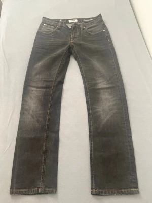 Svarta jeans från Nudie Jeans - Säljer ett par svarta jeans från Nudie Jeans med snyggt slitna detaljer och klassisk femficksmodell. Jeansen har raka ben och kontrastsömmar i orange. Materialet är denim i bomull och passformen är normal. Perfekta för en avslappnad och stilren look.