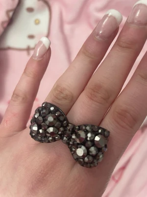 Svart rosettring med glittrande stenar - Cool ring i form av en rosett, täckt med svarta och silvriga glittrande stenar. Ringen har en chunky design som verkligen sticker ut och passar perfekt för dig som gillar statement-smycken. Materialet är metall med dekorerade stenar.