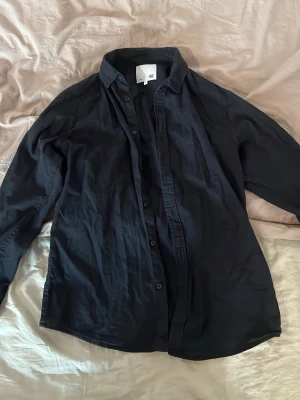 Svart skjorta från 157, storlek M - Klassisk svart skjorta från 157 i storlek M. Skjortan har lång ärm, knappar framtill och en stilren krage. Perfekt för dig som gillar en enkel och tidlös look. Tillverkad i mjukt bomullstyg för skön känsla hela dagen.