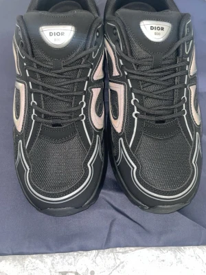 Svarta Dior sneakers med vita detaljer - Säljer ett par svarta Dior sneakers med vita och ljusrosa detaljer. Skorna har en chunky sula, meshpaneler och reflekterande linjer längs ovandelen. Klassisk snörning och logga på plösen. Perfekta för dig som vill ha en statement sneaker från ett lyxigt märke.