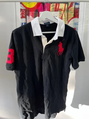 Kortärmad piké från Polo Ralph Lauren - Svart pikétröja från Polo Ralph Lauren med vit krage och knappslå. Stor röd logga på bröstet och röd siffra 3 på ärmen. Klassisk och sportig stil, perfekt för dig som gillar preppy mode. Storlek XL i barn dvs M i senior. 