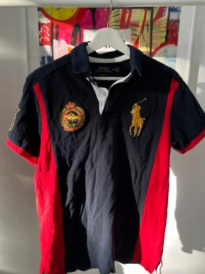 Polo Ralph Lauren svart/röd t-shirt - Snygg svart och röd t-shirt från Polo Ralph Lauren med klassisk pikékrage och stora broderade loggor i guld på bröstet. T-shirten har korta ärmar, vita detaljer vid kragen och är tillverkad i bomull. Perfekt för dig som gillar sportig och stilren design.