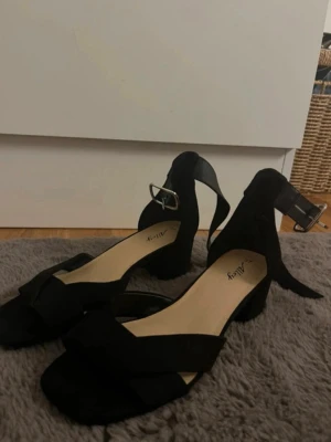 Svarta sandaler med klack från Alley - Stilrena svarta sandaler från Alley med breda remmar i mockaimitation och öppen tå. Matrialet är mocka. Skorna har en stadig blockklack och justerbart spänne runt ankeln. Perfekta för dig som vill ha en klassisk och trendig look med bekväm passform.