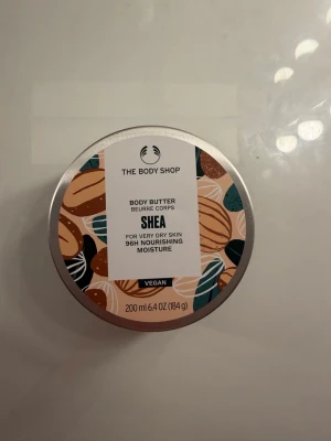 Shea Body Butter från The Body Shop - Shea Body Butter från The Body Shop, 200 ml. Vegansk kroppssmörj med sheasmör, passar extra torr hud. Snygg rund burk med färgglad design i beige, brunt, grönt och vitt.