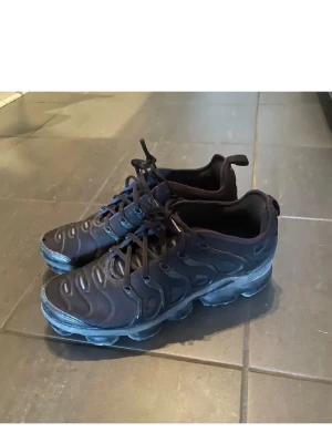 Nike Air Max Plus TN svarta sneakers - Säljer ett par svarta Nike Air Max Plus TN sneakers med vågigt mönster på ovandelen och kraftig sula. Skorna har snörning och är tillverkade i syntetmaterial och mesh. Perfekt för dig som gillar en sportig och cool look. Litet hål längst ut men det synns ej