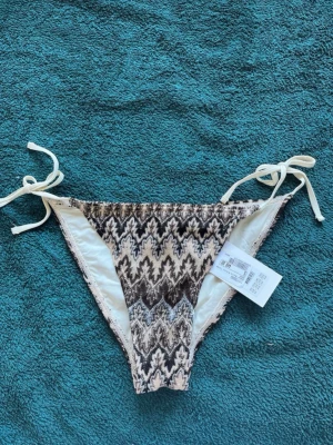 Mönstrad bikiniunderdel med knyt - Säljer en snygg bikiniunderdel med zigzagmönster i svart, beige och grå toner. Underdelen har knyt i sidorna för justerbar passform och är tillverkad i ett mjukt syntetmaterial. Perfekt för stranden eller poolen!