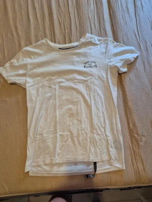 Vit t-shirt med tryck 'Live Life No Regrets' - Vit t-shirt med svart texttryck 'Live Life No Regrets' på bröstet. Klassisk rund halsringning och korta ärmar. Perfekt för en avslappnad och stilren look.