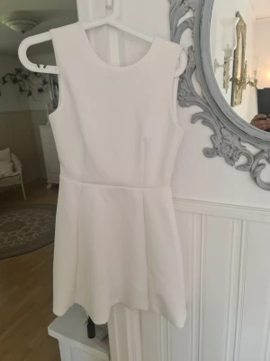 Vit ärmlös kortklänning med öppen rygg - Snygg vit kortklänning med ärmlös design och djup öppen rygg. Klänningen har en enkel, stilren look och lätt utsvängd kjol. Perfekt för dig som gillar minimalistisk stil och vill sticka ut med en öppen rygg.