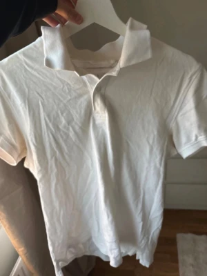 Vit pikétröja från Kappahl - Klassisk vit pikétröja från Kappahl med krage och korta ärmar. Tröjan är i bomull och har en enkel, stilren design med knappar vid halsen. Perfekt för en clean och fräsch look.