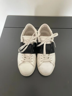 Vit-svarta sneakers från Valentino - Snygga vita sneakers från Valentino med en bred svart läderdetalj över mitten. Använd men fortfarande i mycket bra skick. Inga större defekter än att man ser på bilderna. Det är bara att buda!!