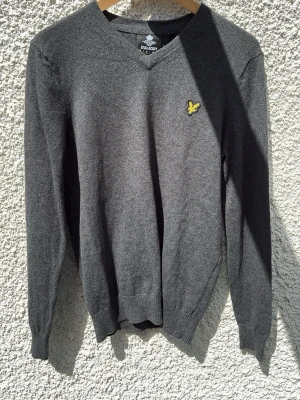 Grå stickad tröja från Lyle & Scott - Mörkgrå stickad tröja från Lyle & Scott med V-ringning och den klassiska gula loggan broderad på bröstet. Tröjan har långa ärmar och ribbade muddar vid ärmslut och nederkant. Perfekt för lager-på-lager och stilren look.