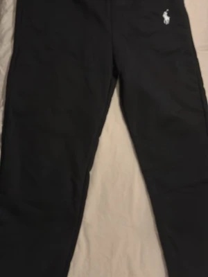 Svarta mjukisbyxor från Polo Ralph Lauren - Svarta mjukisbyxor från Polo Ralph Lauren med klassisk vit logga broderad på höften. Byxorna har resår i midjan och är tillverkade i mjukt material som känns skönt mot huden. Perfekta för en avslappnad och stilren look.