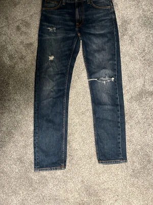 Mörkblå slitna jeans från Nudie - Säljer ett par mörkblå jeans från Nudie med snygga slitningar och hål på benen. Jeansen har klassisk femficksdesign, orangea sömmar och raka ben. Perfekta för en avslappnad och trendig stil.