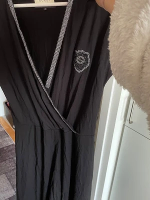Svart omlottklänning med strassdetaljer - Stor i storlek! Som en M. Svart omlottklänning från Cecil med v-ringning och strassdetaljer längs kanten och vid bröstet. Klänningen har korta ärmar och dekorativ kant nertill. Perfekt för dig som gillar en elegant och stilren look.