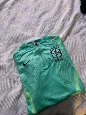 Nike Brasil träningströja turkos - Snygg turkos träningströja från Nike med Brasil-logga och CBF-märke på bröstet. Tröjan har halv dragkedja framtill och tunna vita linjer som design. Perfekt för fotbollsträning eller sportiga aktiviteter.