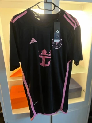 Svart Inter Miami matchtröja Adidas - Snygg svart matchtröja från Adidas för Inter Miami CF med rosa detaljer och klubbmärke på bröstet. Tröjan har rund hals, korta ärmar och är tillverkad i ett lätt, ventilerande material. Slim fit-modell som passar perfekt till träning eller match.
