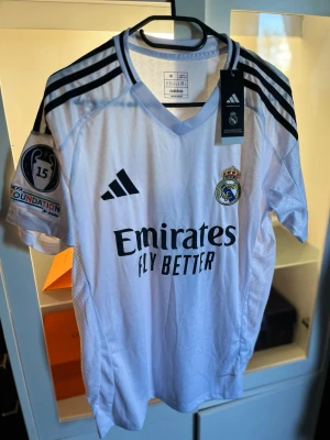 Real Madrid matchtröja Adidas M - Vit Real Madrid matchtröja från Adidas med svarta detaljer och klubbmärke på bröstet. Tröjan har V-ringning, broderad logga, Champions League-märke på ärmen och texten 'Emirates Fly Better' framtill. Slim fit-modell i lätt och ventilerande material.