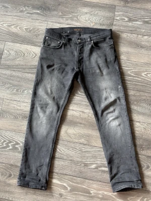 Grim tim Nudie Jeans - Snygga grå slim fit jeans från Nudie Jeans med klassisk femficksdesign och diskreta slitningar. Jeansen har knappgylf och är tillverkade i bomull med en skön stretch. Perfekta för dig som gillar en avslappnad men stilren look.