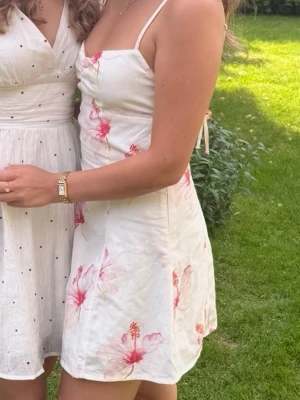 Peppermayo klänning med blommor  - Söt vit kortklänning med rosa blommönster och smala axelband. Klänningen har en figurnära passform och är perfekt för varma dagar. Lätt och luftig känsla, med en feminin och somrig look. Den är även öppen i ryggen med knyten 