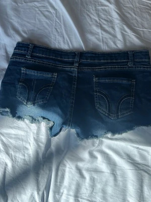Blå slitna jeansshorts från Hollister - Snygga blå jeansshorts från Hollister med slitna detaljer och fransig kant. Klassisk femficksmodell med låg midja och tydliga kontrastsömmar. Perfekta för sommardagar när du vill ha en avslappnad och trendig look.