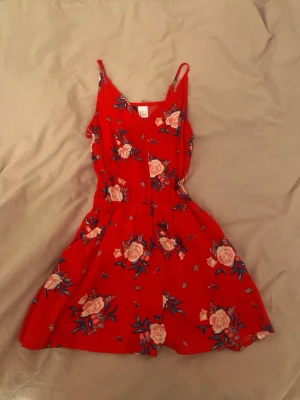 Röd blommig jumpsuit med smala axelband - Superfin röd jumpsuit med rosa och vita blommor samt smala axelband. Modellen har markerad midja och korta ben, perfekt för sommardagar. Materialet känns lätt och luftigt, vilket gör den skön att bära. Snygg och trendig design som sticker ut.