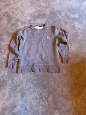 Grå sweatshirt från Acne Studios - Säljer en stilren grå sweatshirt från Acne Studios med klassisk rund halsringning och diskret logopatch på bröstet. Tröjan är i mjuk bomullsblandning och har ribbade muddar vid ärmslut och nederkant. Perfekt för en avslappnad look. Ny pris 2000kr när den fanns att köpa ny.