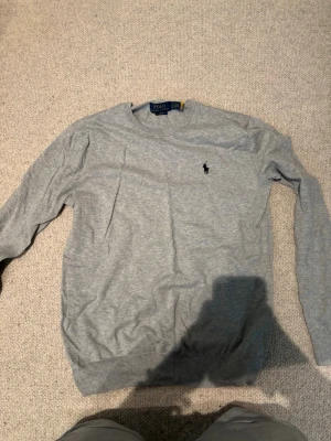 Grå tröja från Polo Ralph Lauren - Snygg grå tröja från Polo Ralph Lauren med klassisk rund halsringning och den ikoniska lilla broderade loggan på bröstet. Tröjan har långa ärmar och ribbade muddar vid ärmslut och nederkant. Perfekt till jeans eller chinos.