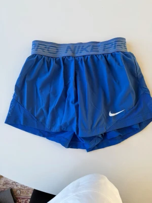 Blå shorts från Nike - Snygga blå shorts från Nike med bred elastisk midjeresår där det står 'NIKE PRO'. Shortsen har en vit Nike-logga framtill och är tillverkade i ett lätt och ventilerande material, perfekt för träning och löpning. Shortsen har undershirts.