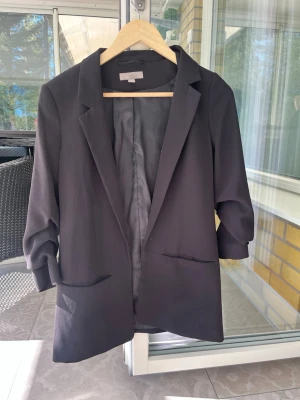 Svart kavaj med klassisk passform - Stilren svart kavaj med klassisk krage och två fickor framtill. Kavajen har långa ärmar och är helfodrad på insidan. Perfekt för en snygg och enkel look. Passar till många olika outfits.