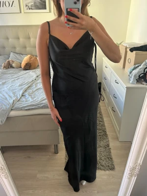 Svart balklänning från ASOS DESIGN - Elegant svart balklänning i satin med slit och tre dekorativa rosetter längs ena sidan som ger en exklusiv och feminin detalj ✨  Perfekt till bal, gala, bröllop eller andra festliga tillfällen 🖤  Klänningen har ett vackert fall och känns lyxig i materialet. Klänningen är helt ny och orginalförpackning medföljer.  Jag är 178 cm lång och bär vanligtvis M och den går nästan ned till fotknölarna, perfekt längd för klackar 👠 