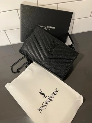  YSL väska - Säljer en svart handväska från Yves Saint Laurent med klassisk YSL-logga framtill. Väskan är tillverkad i strukturerat läder med chevronmönster och har en elegant kedjerem i svart metall. Kommer med original dustbag och låda. Säljs så snabbt som möjligt !! Pris kan diskuteras!! 