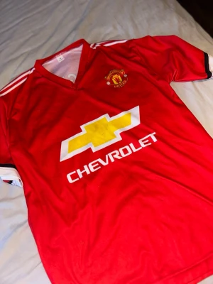 Manchester United Pogba matchtröja - Röd Manchester United matchtröja med Paul Pogba’s namn på ryggen från Adidas med klubbmärke på bröstet och Chevrolet-logga framtill. Tröjan har vita detaljer på axlarna och svart/vita muddar vid ärmarna. Perfekt för fotbollsfans som vill visa sitt stöd på läktaren eller planen. Pris kan diskuteras.