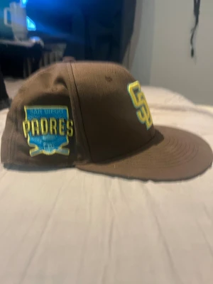 San Diego Padres brun keps - Säljer en brun keps med broderad SD-logga i gult och blått framtill samt San Diego Padres-märke på sidan. Klassisk flat brim och justerbar passform. Perfekt för dig som gillar baseball eller vill ha en sportig stil.