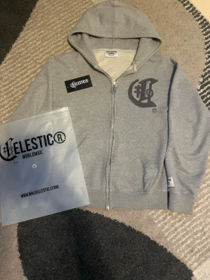 Grå zip hoodie från Celestic - Snygg grå zip hoodie från Celestic med stor broderad logga på bröstet och matchande dragkedja. Hoodien har huva, två fickor fram och ribbade muddar. Perfekt för en chill och streetwear-inspirerad look.