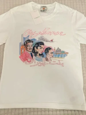 Vit Casablanca t-shirt med tryck - Vit t-shirt från Casablanca med färgglatt tryck framtill som visar personer, byggnader och ett flygplan. T-shirten har rund hals och korta ärmar. Perfekt för dig som gillar unika och konstnärliga motiv.