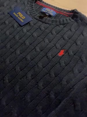 Mörkblå kabelstickad tröja Polo Ralph Lauren - Snygg mörkblå kabelstickad tröja från Polo Ralph Lauren med klassisk röd broderad logga på bröstet. Tröjan har rund halsringning och ribbade muddar. Perfekt för dig som gillar tidlös och stilren design. BARN STORLEK PASSAR XS