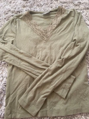 Olivgrön långärmad topp Gina Tricot - Olivgrön långärmad topp från Gina Tricot med v-ringning och dekorativ spets runt halsen. Tillverkad i mjukt material som känns skönt mot huden. Perfekt för dig som gillar enkla men snygga detaljer.
