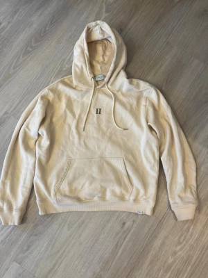 Beige hoodie med broderad detalj - Stilren beige hoodie från Les Deux med broderad svart 'II' på bröstet. Klassisk känguruficka, ribbade muddar och justerbar huva med dragsko. Perfekt för lager-på-lager och chill vardagsstil.