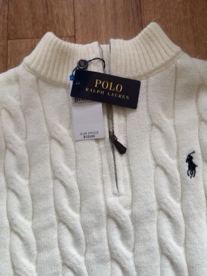 Vit kabelstickad tröja Polo Ralph Lauren - Vit kabelstickad tröja från Polo Ralph Lauren med halv dragkedja och hög krage. Tröjan har klassisk kabelstickning och den ikoniska broderade loggan på bröstet. Perfekt för lager-på-lager och snygg till jeans.