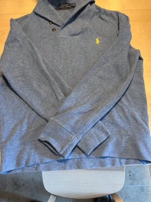 Blå långärmad tröja från Polo Ralph Lauren - Snygg blå långärmad tröja från Polo Ralph Lauren med klassisk krage och tre knappar vid halsen. Passar som en S Tröjan har gul broderad logga på bröstet och är tillverkad i mjukt material. Pris kan diskuteras vid snabbt köp