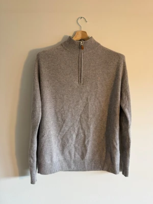Morris Stockholm Quarter Zip - Grey Wool - Morris Stockholm quarter-zip i 100% lambswool – stl L, grå melange. Armbågslapper i matchande material, YKK-zipp med läderdetalj och fleur-de-lis-emblem. Klassisk prep-estetik med bra materialvikt. Skick: som ny