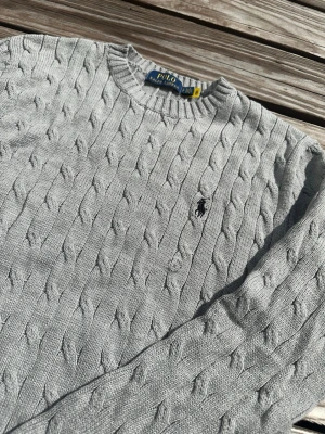 Ljusgrå kabelstickad tröja Polo Ralph Lauren - Kabelstickad ljusgrå tröja från Polo Ralph Lauren med rund halsringning och broderad logga på bröstet. Tröjan har långa ärmar och ribbade muddar vid ärmslut och nederkant. Perfekt för lager-på-lager och klassisk stil.