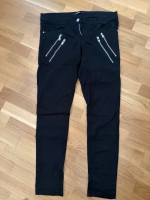 Svarta jeans med dragkedjor från Bik Bok - Säljer ett par svarta skinny jeans från Bik Bok med fyra coola dragkedjor framtill. Jeansen har normal midja, bälteshällor och knappstängning. Materialet är stretchigt och bekvämt, perfekt för en trendig look.