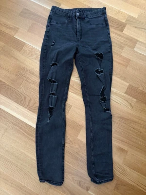 Svarta ripped jeans från Divided - Snygga svarta jeans från Divided med flera slitningar och hål på benen för en trendig look. Modellen är slim fit med hög midja och klassisk femficksdesign. Perfekta för dig som gillar en edgy stil.
