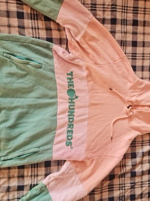 Pastellfärgad hoodie från The Hundreds - Säljer en snygg hoodie från The Hundreds i pastellrosa och mintgrönt. Hoodien har huva med dragsko, magficka med dragkedja och stort tryck på bröstet. Perfekt för dig som gillar streetwear och vill sticka ut med färg.
