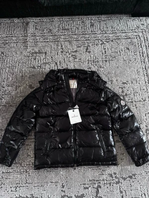  Moncler jacka  - Pris kan diskuteras privat. Snygg svart dunjacka från Moncler med glansig finish och huva. Jackan har dragkedja framtill, två fickor med dragkedja och Moncler-logga på ärmen. Perfekt för kalla vinterdagar och riktigt trendig stil.