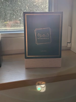 Lattafa Khamrah Eau de Parfum 100ml fast pris - Lattafa Khamrah Eau de Parfum kommer i en elegant, fyrkantig glasflaska med detaljer i guld och ett snyggt mönster. Förpackningen är svart med gulddetaljer och en träfärgad botten. Flaskan rymmer 100 ml och har en lyxig känsla som passar parfymälskare. Helt ny skit bra pris 