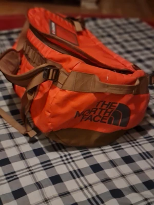 Orange resväska från The North Face - Säljer en snygg och rymlig resväska från The North Face i orange och brunt. Väskan har robusta remmar, flera fack och stor logga på sidan. Perfekt för resor eller träning, tillverkad i slitstarkt material och med praktiska detaljer.