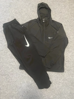 Svart Nike vindjacka med huva - Svart vindjacka från Nike med huva och dragkedja. Jackan har reflekterande detaljer och Nike-logga på bröstet. Perfekt för löpning eller utomhusaktiviteter. Tillverkad i lätt och smidigt material som skyddar mot vind.